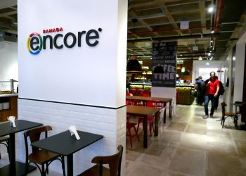 Entrada do Hotel Ramada Encore Tiradentes. É possível ver mesas em uma área de alimentação, uma recepção ao fundo e dois homens em pé, no corredor à direita