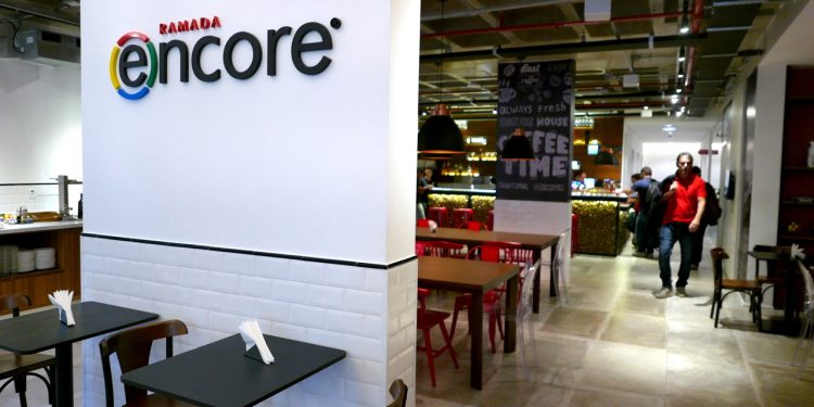 Entrada do Hotel Ramada Encore Tiradentes. É possível ver mesas em uma área de alimentação, uma recepção ao fundo e dois homens em pé, no corredor à direita